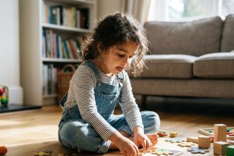 Petite fille de 5 ans concentrée sur un puzzle en bois coloré posé sur le plancher d'un salon chaleureux
