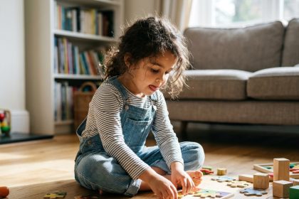 Petite fille de 5 ans concentrée sur un puzzle en bois coloré posé sur le plancher d'un salon chaleureux