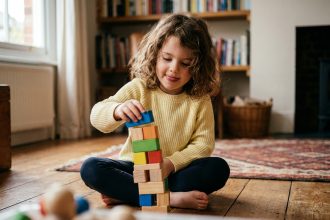 Petite fille concentrée qui empile des cubes en bois colorés sur un parquet en bois, jouet éducatif de motricité fine